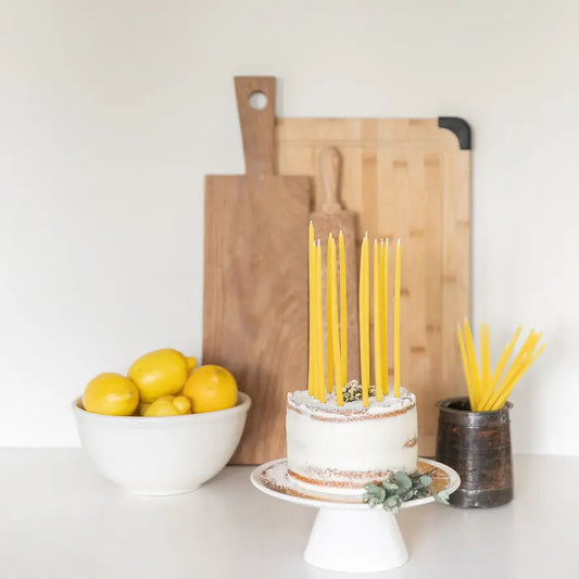 Unlit thin beeswax candles on a frosted cake on a white stand beside lemons and cutting boards.