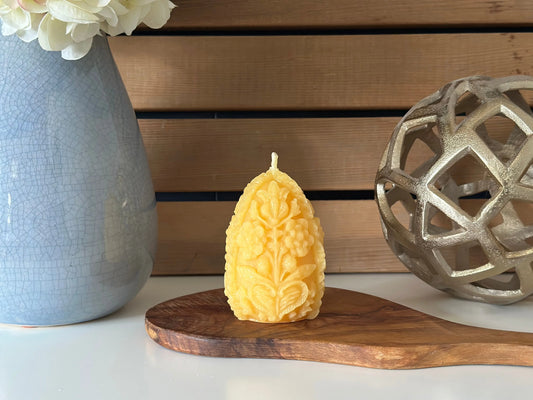 Carved floral egg candle on an olive wood board with a blue vase and a metal sphere behind.