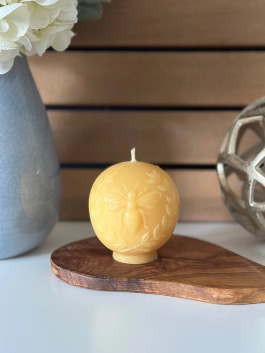 Bee globe candle on an olive wood board in front of a grey vase and wood slat wall.