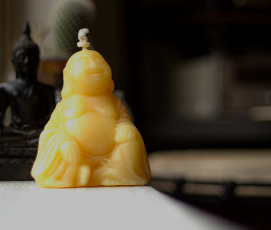 Laughing Buddha beeswax candle on a ledge with a dark Buddha figure blurred behind it.