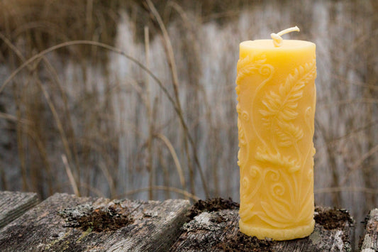 Tall pillar candle with swirling fern relief standing on weathered wood in front of dry grasses.