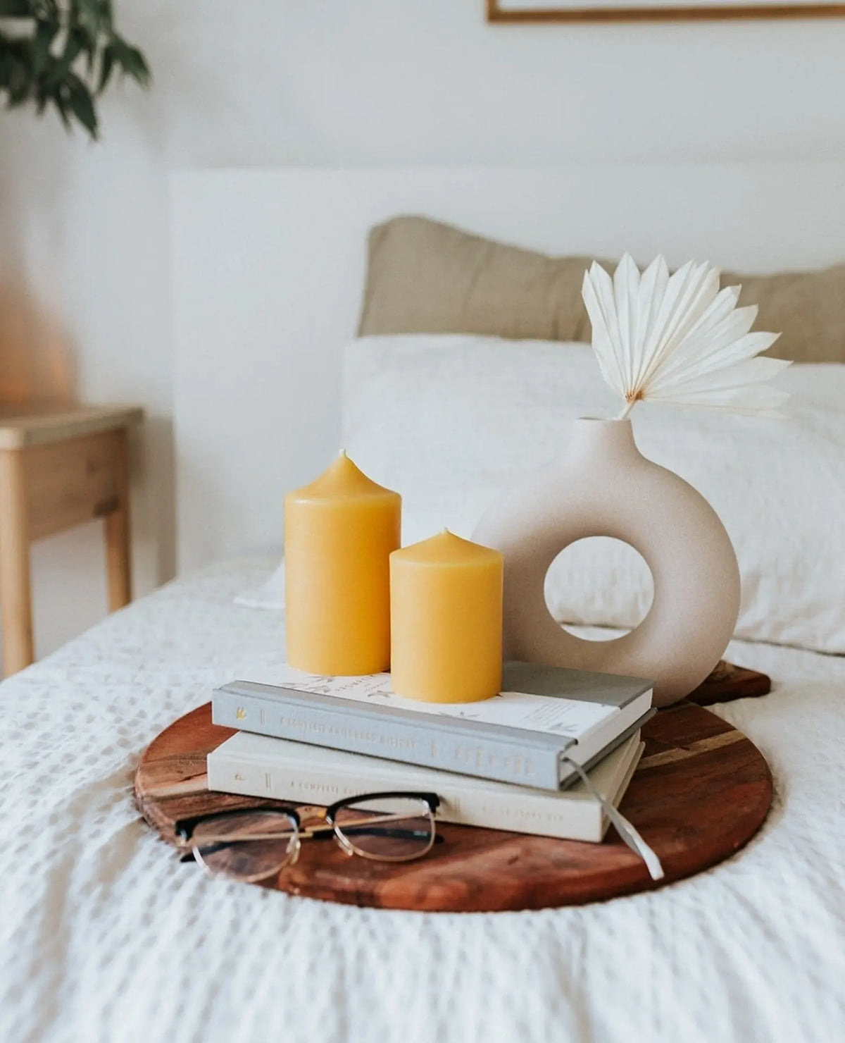 Two bell-top pillars on a round wood tray with books and glasses on a white bed.