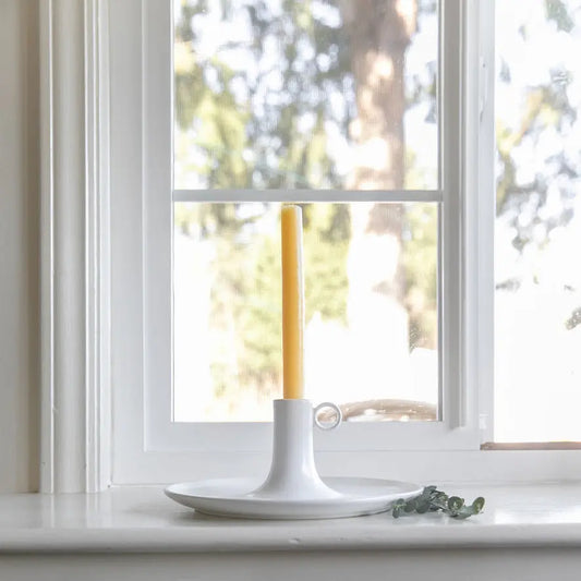 Unlit taper candle in a white chamberstick on a windowsill with trees outside.