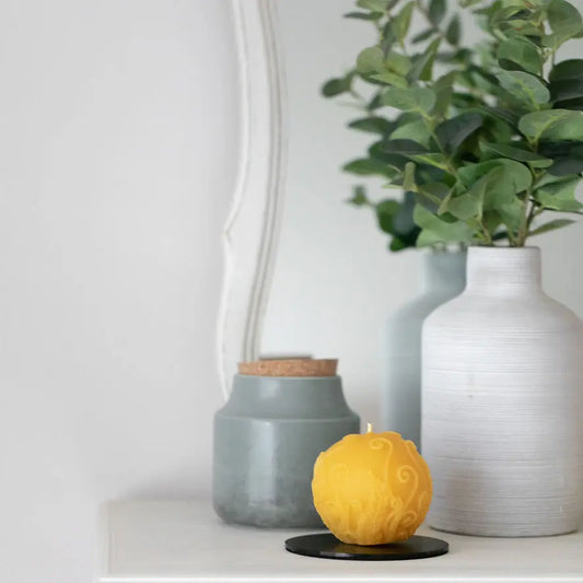 Golden fern-swirl globe candle on a black coaster on a white shelf beside grey vases.