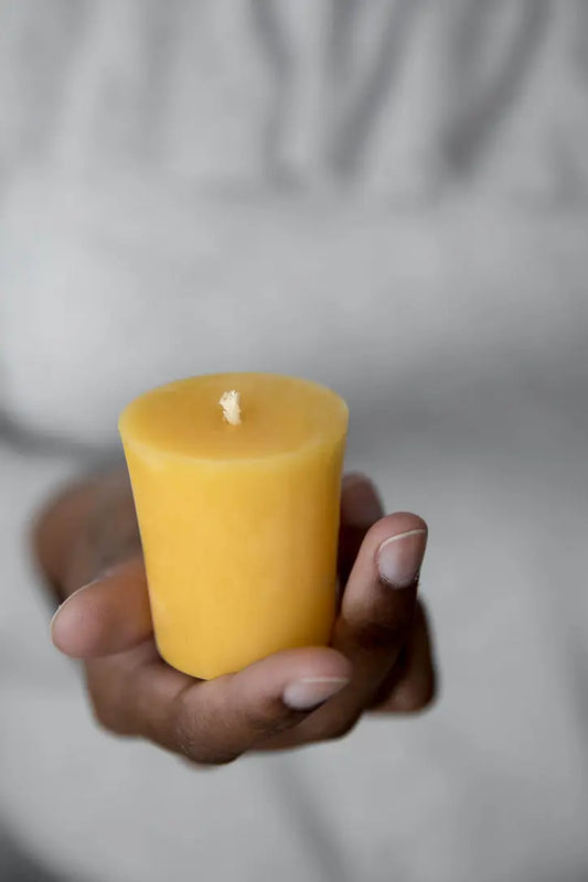 Hand holding a single golden beeswax votive candle in front of a grey sweater.