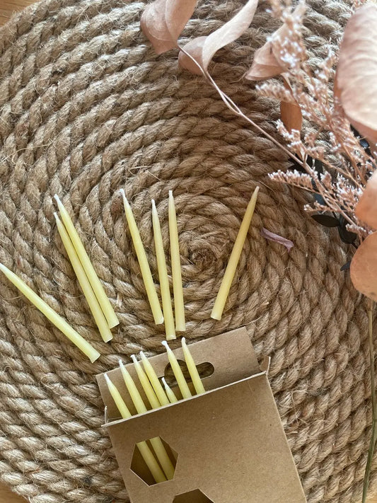 Thin birthday candles spilling from a kraft box onto a coiled rope mat with dried flowers.