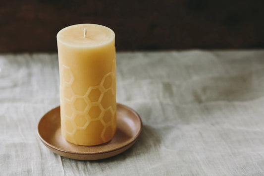 Tall honeycomb-textured pillar candle in pale honey colour on a wooden saucer over grey linen.