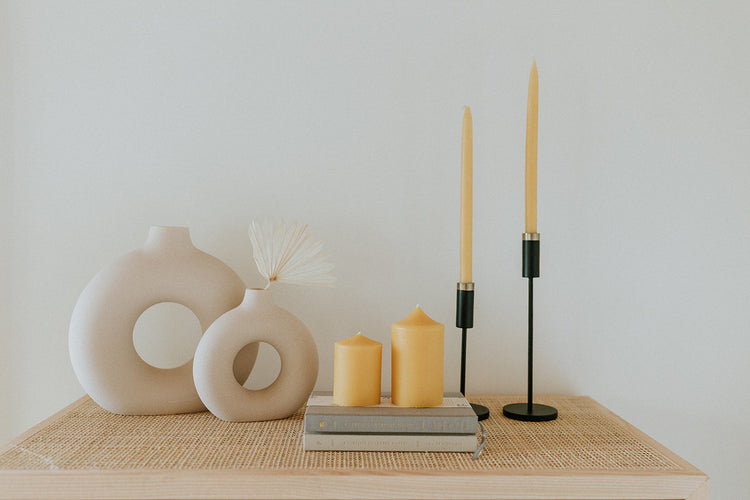 Two beeswax pillars on stacked books with two tall hand-dipped tapers in black candlesticks and cream ring vases on a woven table